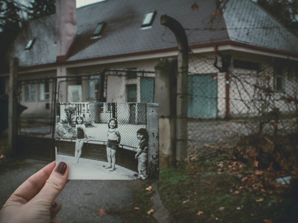 A woman's hand holds a black and white snapshot (of three girls leaning on a fence in front of a well groomed yard of a house) in front of what appears to be the same house at a  later time when the yard and fence are  overgrown and windswept. 